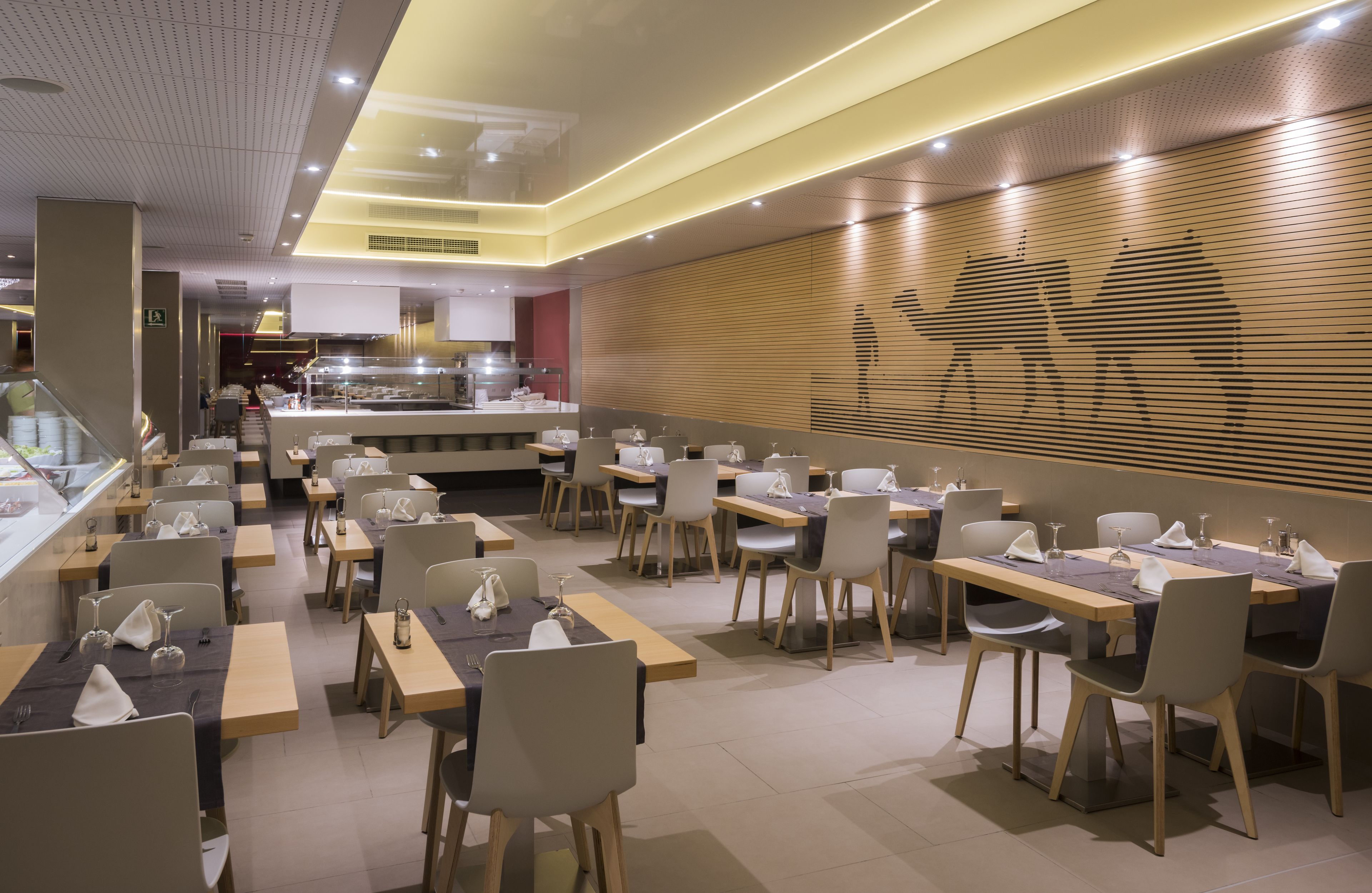 Um restaurante moderno com várias mesas e cadeiras, área de cozinha ao fundo e parede decorativa com design em silhueta.