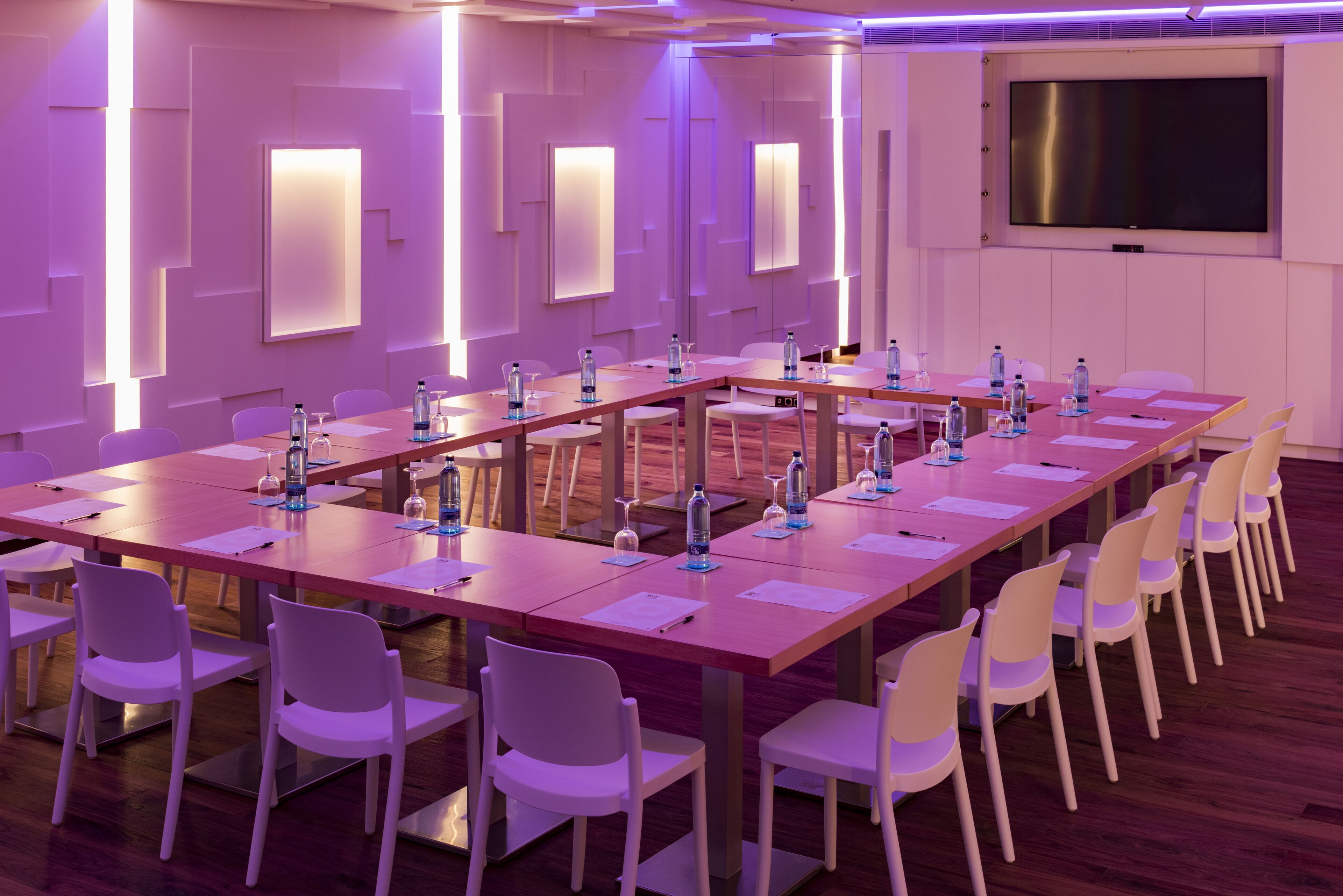Un salón de conferencias con largas mesas, sillas blancas, botellas de agua y una pantalla grande.