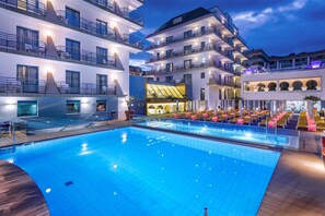 A swimming pool with a wooden deck, surrounded by multi-story buildings with balconies and outdoor seating areas. - Hotel Alhambra (Santa Susanna)