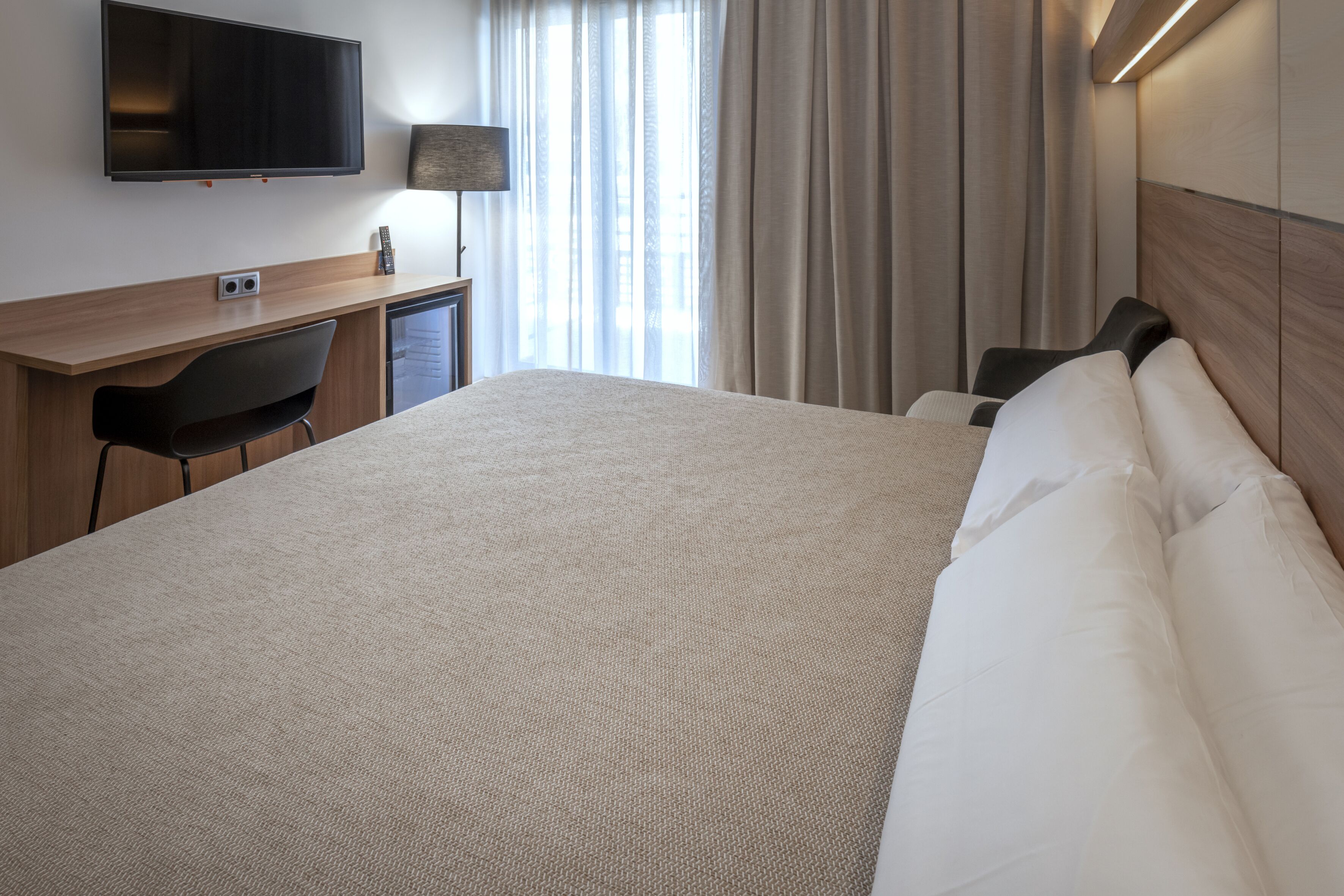 Habitación de hotel con una cama grande, un escritorio con silla, un televisor montado en la pared y una ventana con cortinas.