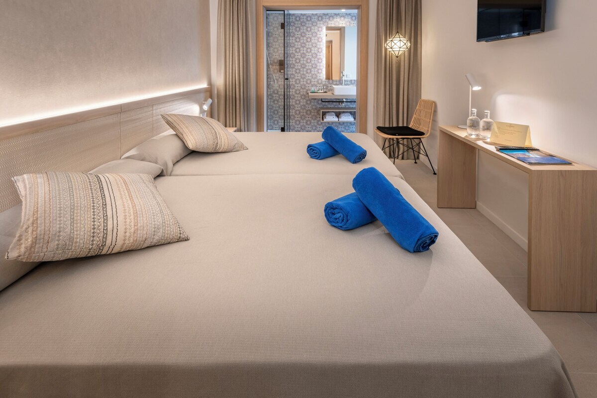 A hotel room with a large bed, two rolled towels, a desk with a lamp, and a mirror.