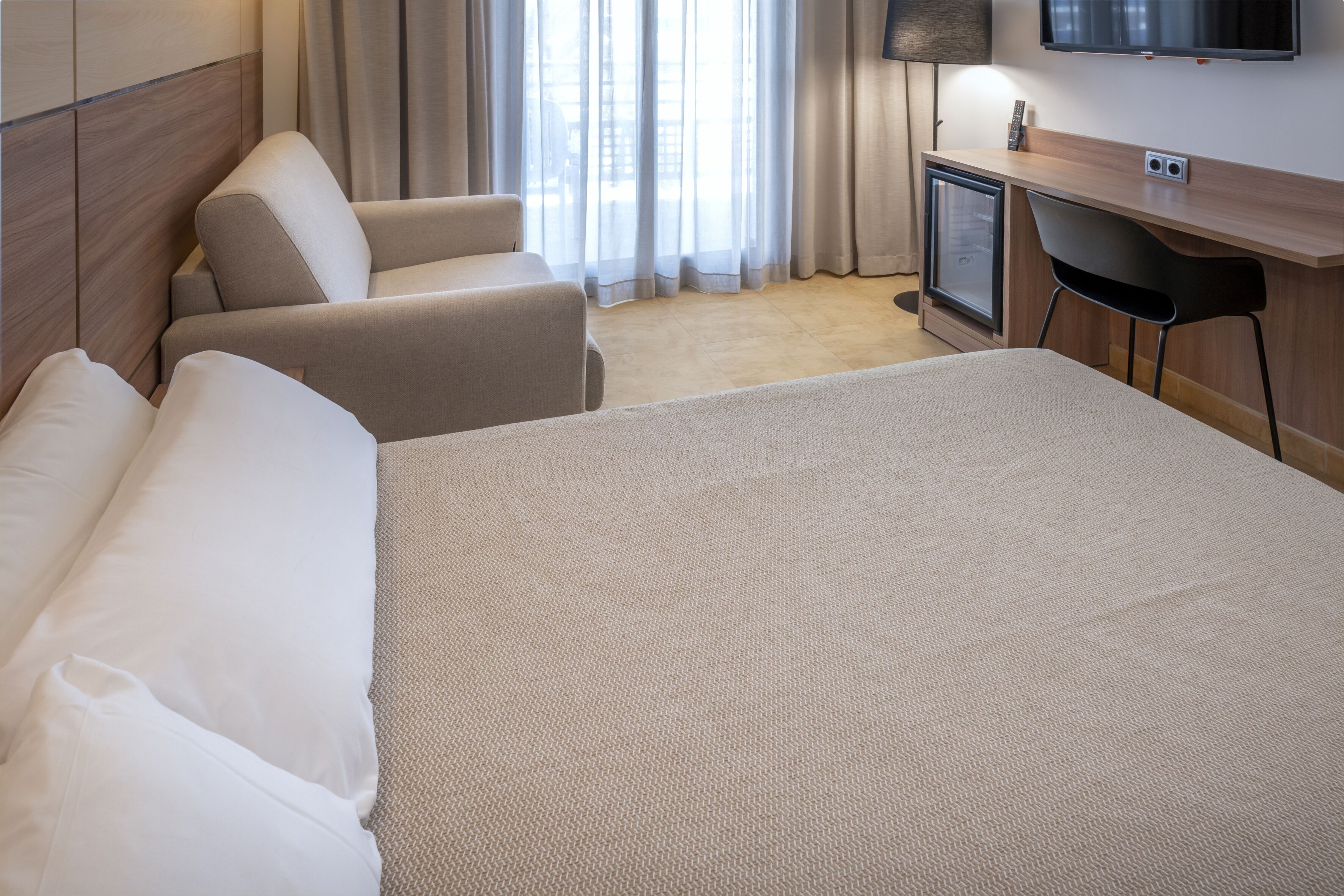A hotel room with a large bed, a sofa, a desk with a TV, and a window with curtains.