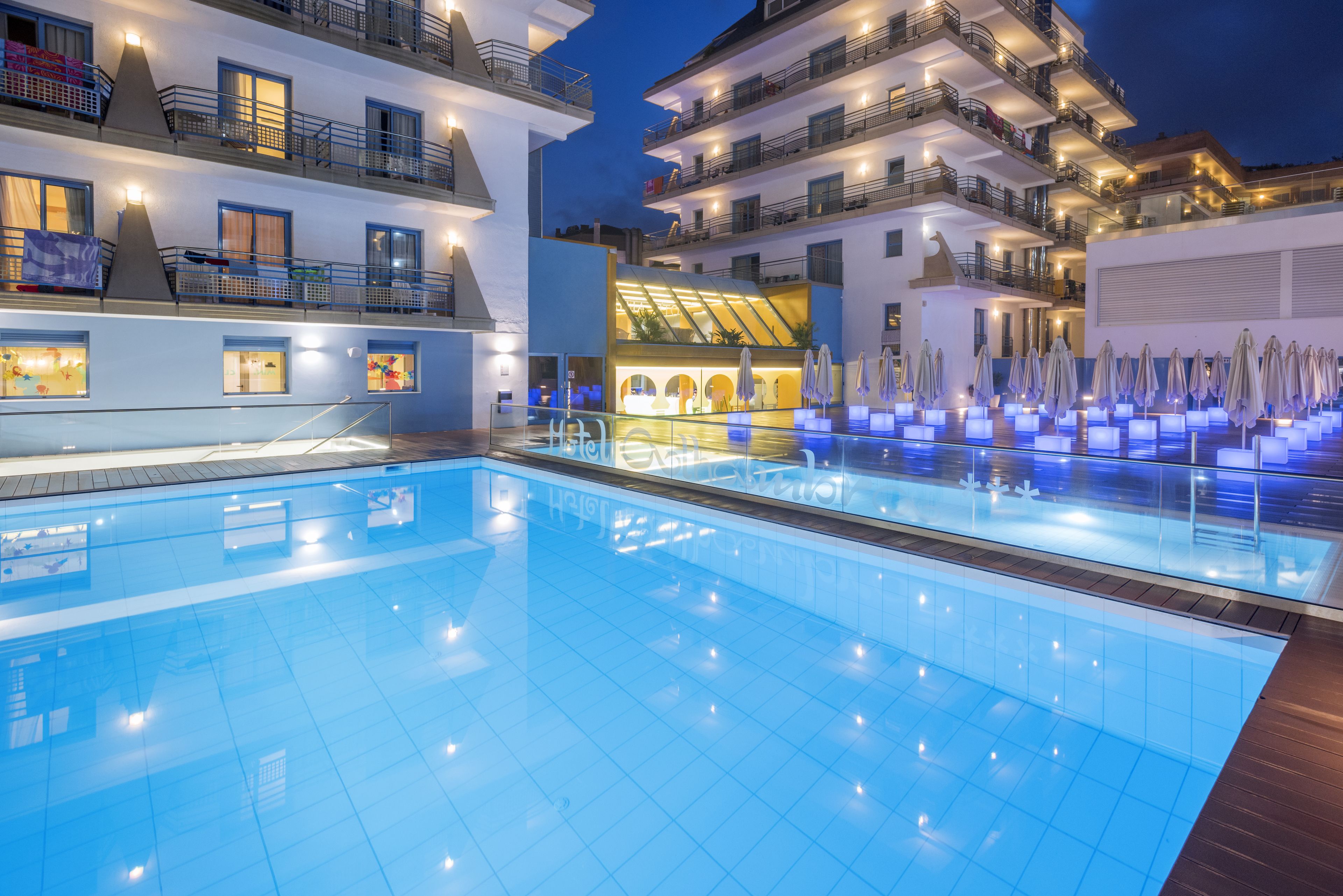 A swimming pool with a glass barrier, adjacent to a building with multiple balconies and outdoor seating areas.