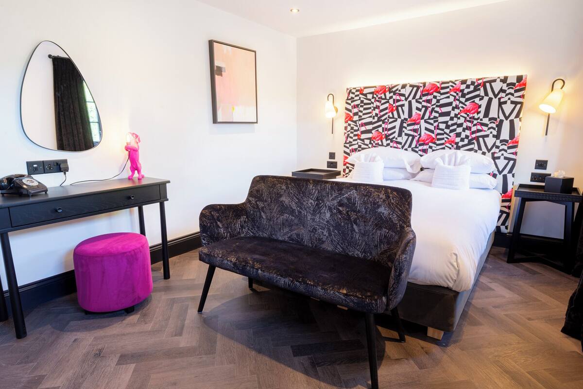 Ett hotellrum med en säng, ett skrivbord, en stol, en spegel och en vägg med ett geometriskt mönster.