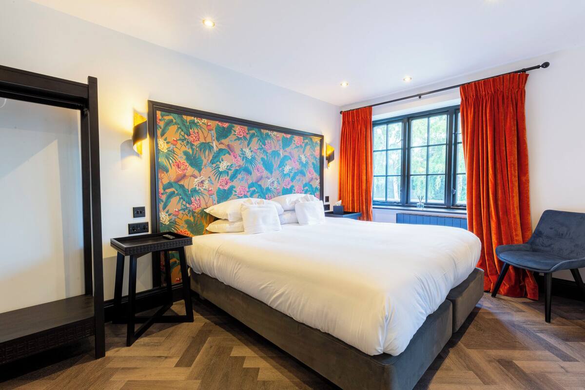 Ett hotellrum med en stor säng, ett skrivbord, en stol, ett fönster med gardiner och en vägg med ett färgstarkt blommönster.