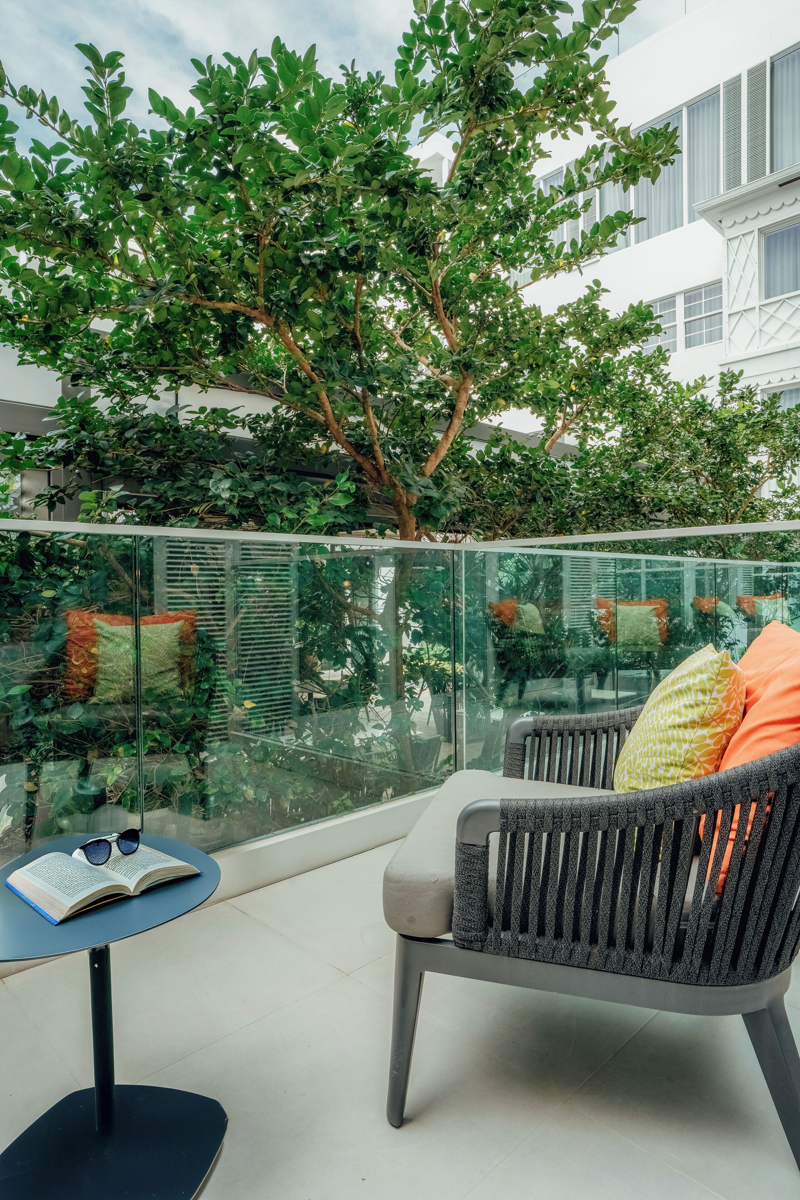 A glass-enclosed balcony with a chair and a small table, a tree, and a building in the background.