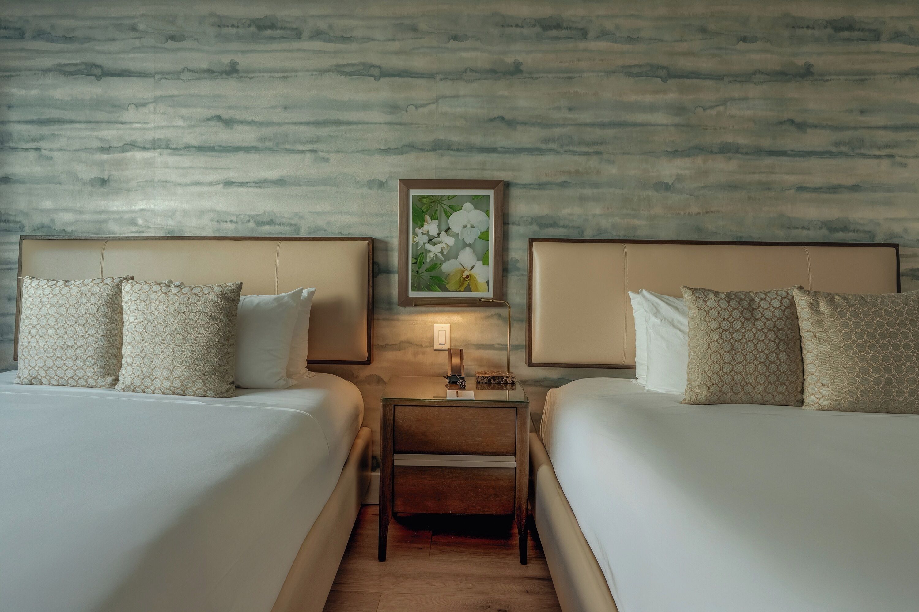 Una habitación de hotel con dos camas, una mesita de noche, una obra de arte floral enmarco y una pared con textura.