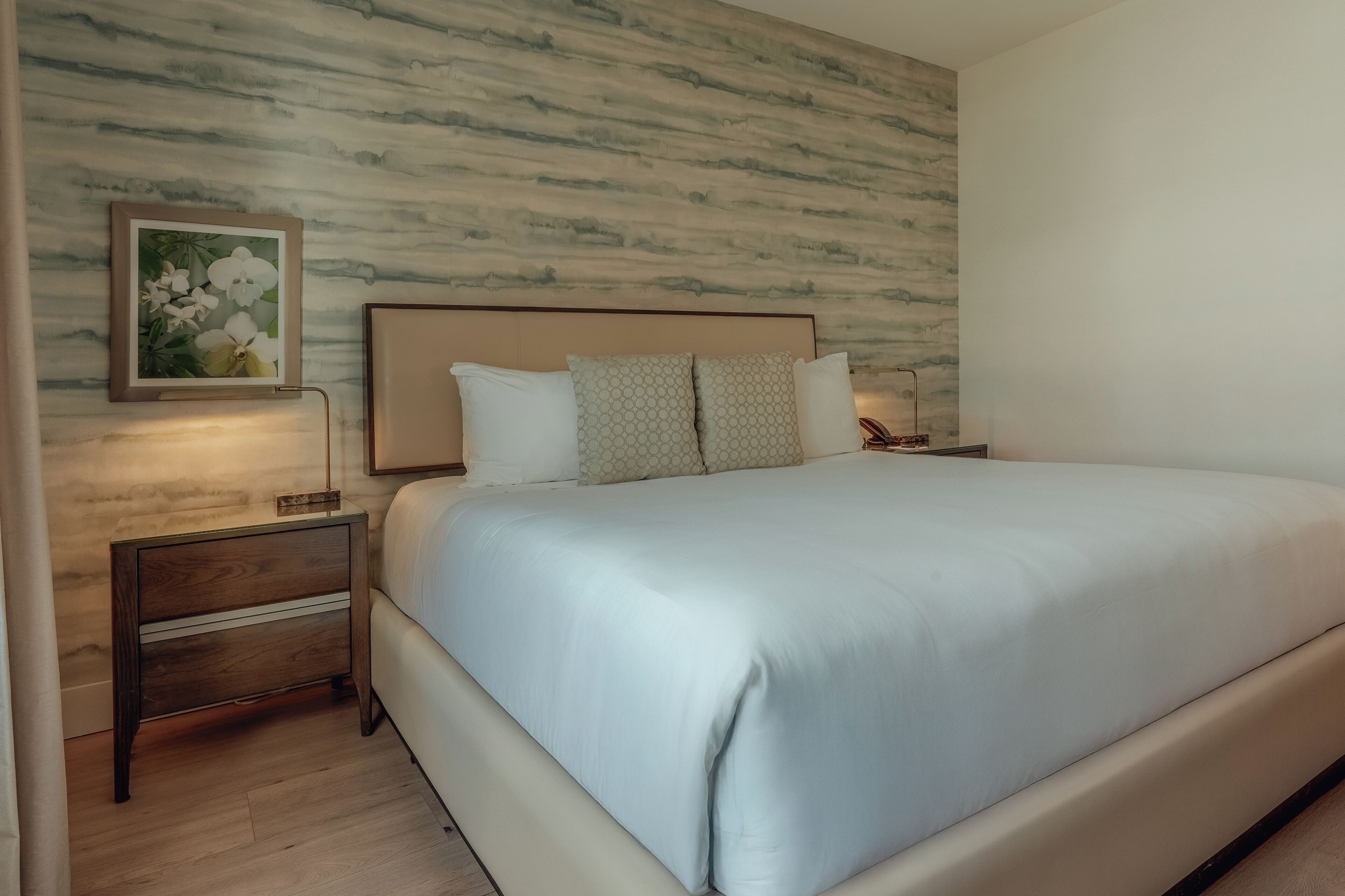 A neatly made bed with a headboard, a bedside table with a lamp, and a framed floral artwork on the wall.