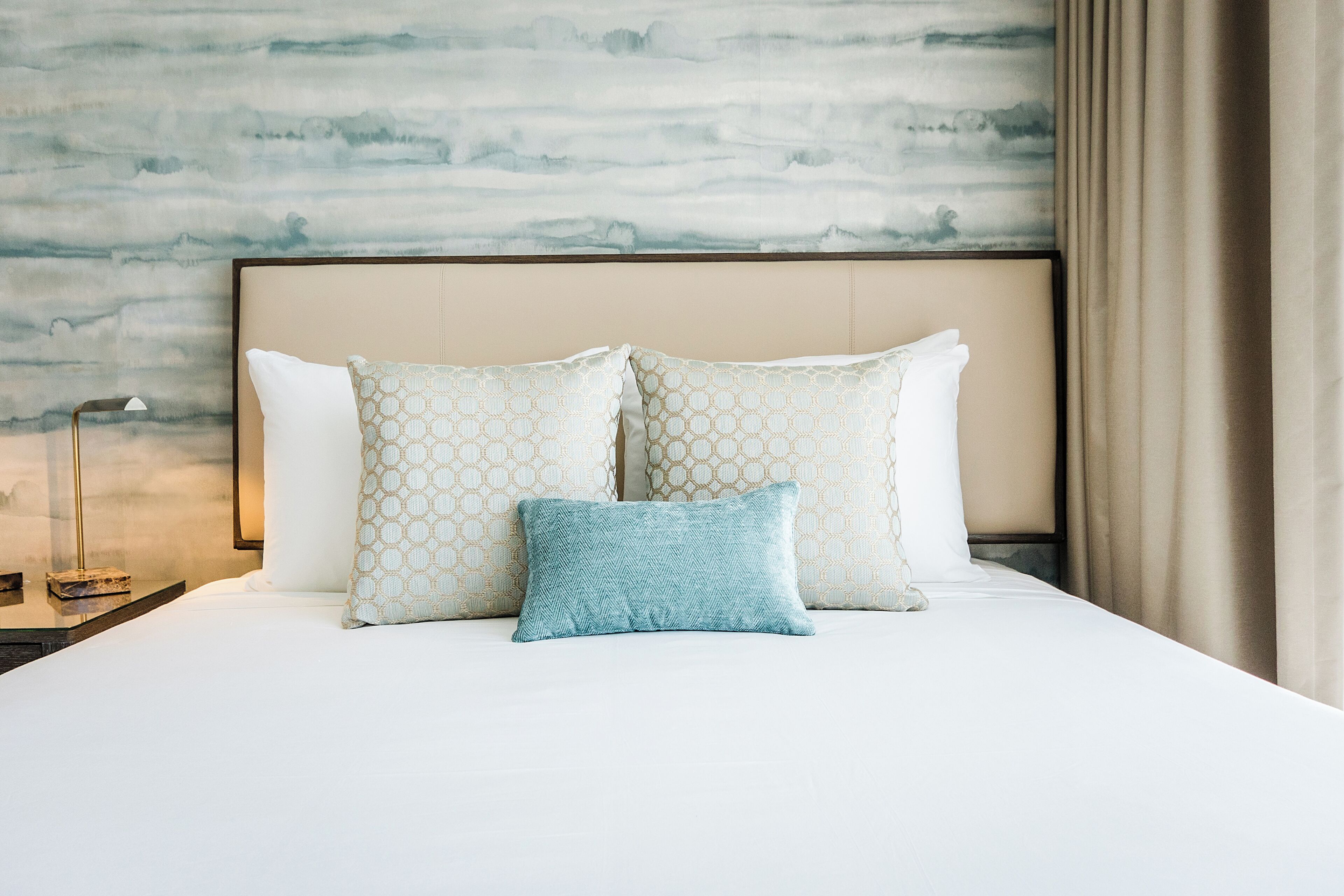 Una cama bien hecha con almohadones estampados, una lámpara de noche y una pared con un mural texturizado.