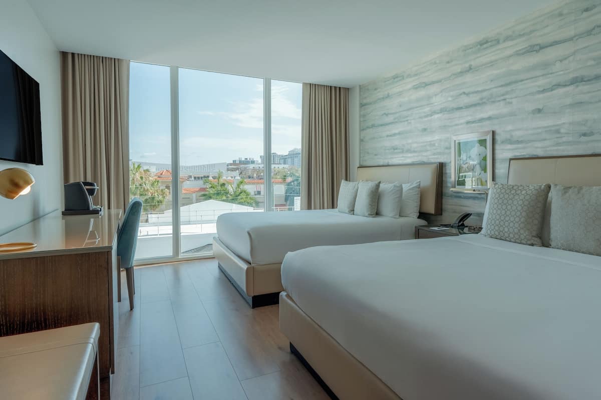 A hotel room with two beds, a desk, a chair, a TV, and a large window with a city view.