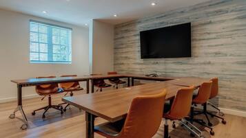 A conference room with a long table, several chairs, a mounted TV, and a window with blinds.