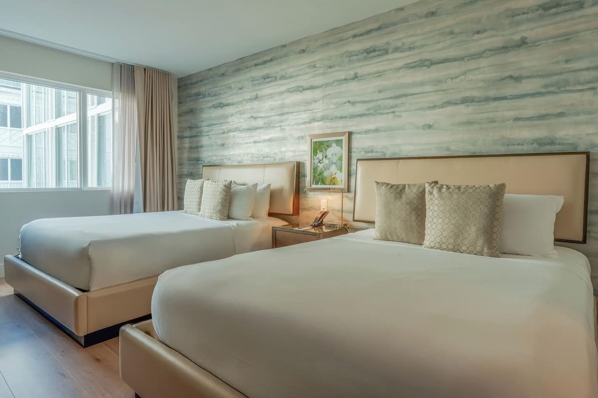 A hotel room with two beds, a nightstand with a lamp, a framed picture on the wall, and a window with curtains.