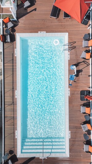 An aerial view of a pool with a wooden deck surrounding it, equipped with lounge chairs and umbrellas.