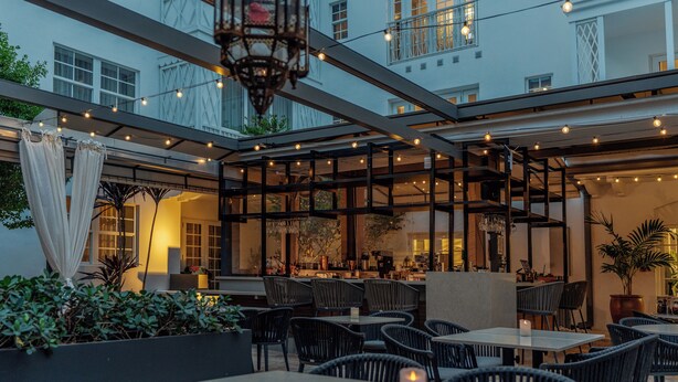 Un espacio de comedor al aire libre con mesas y sillas, luces colgantes y vista al interior del edificio.