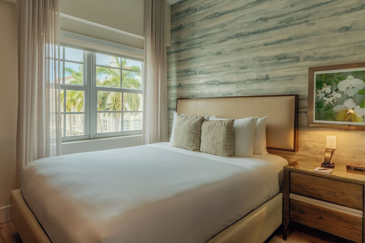 A neatly made bed with a headboard, a nightstand with a lamp, and a window with curtains.