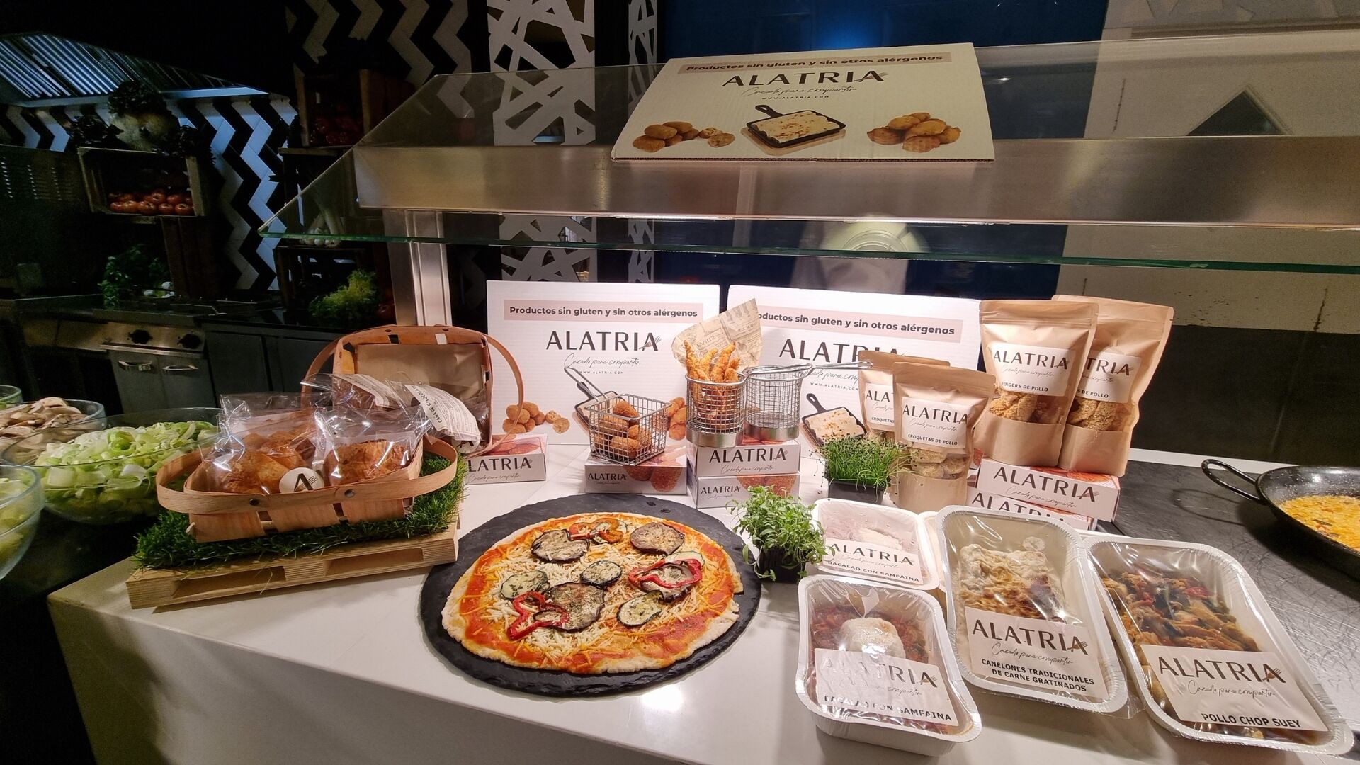 Une table de buffet proposant divers produits alimentaires de la marque ALATRIA, notamment des pizzas, des paniers de pain et des collations emballées.