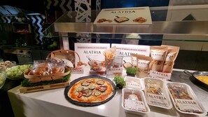Een buffettafel met diverse ALATRIA gerechten, waaronder pizza, broodmandjes en verpakte snacks.