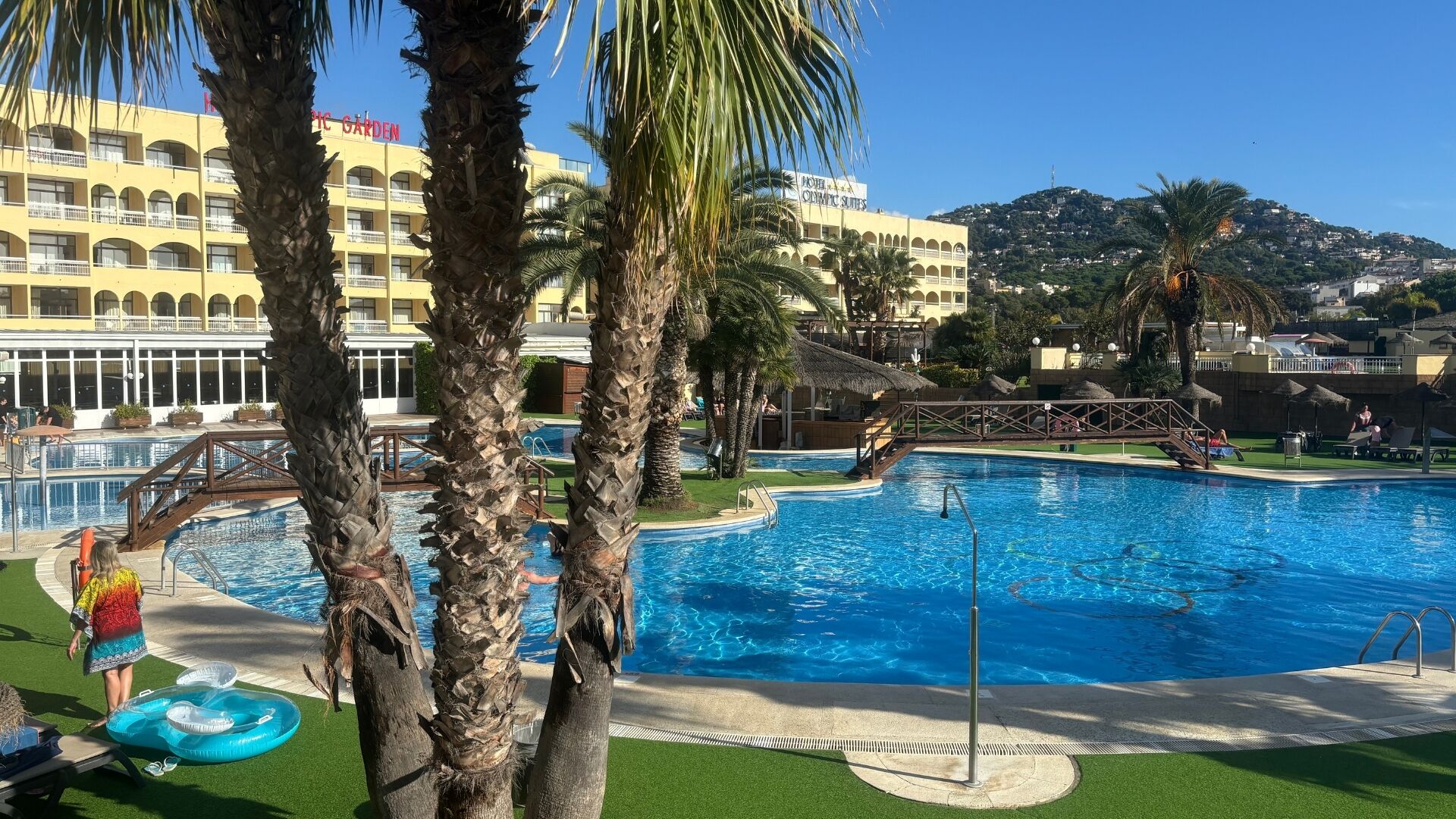 A pool area with palm trees, a bridge, and a hotel building in the background.