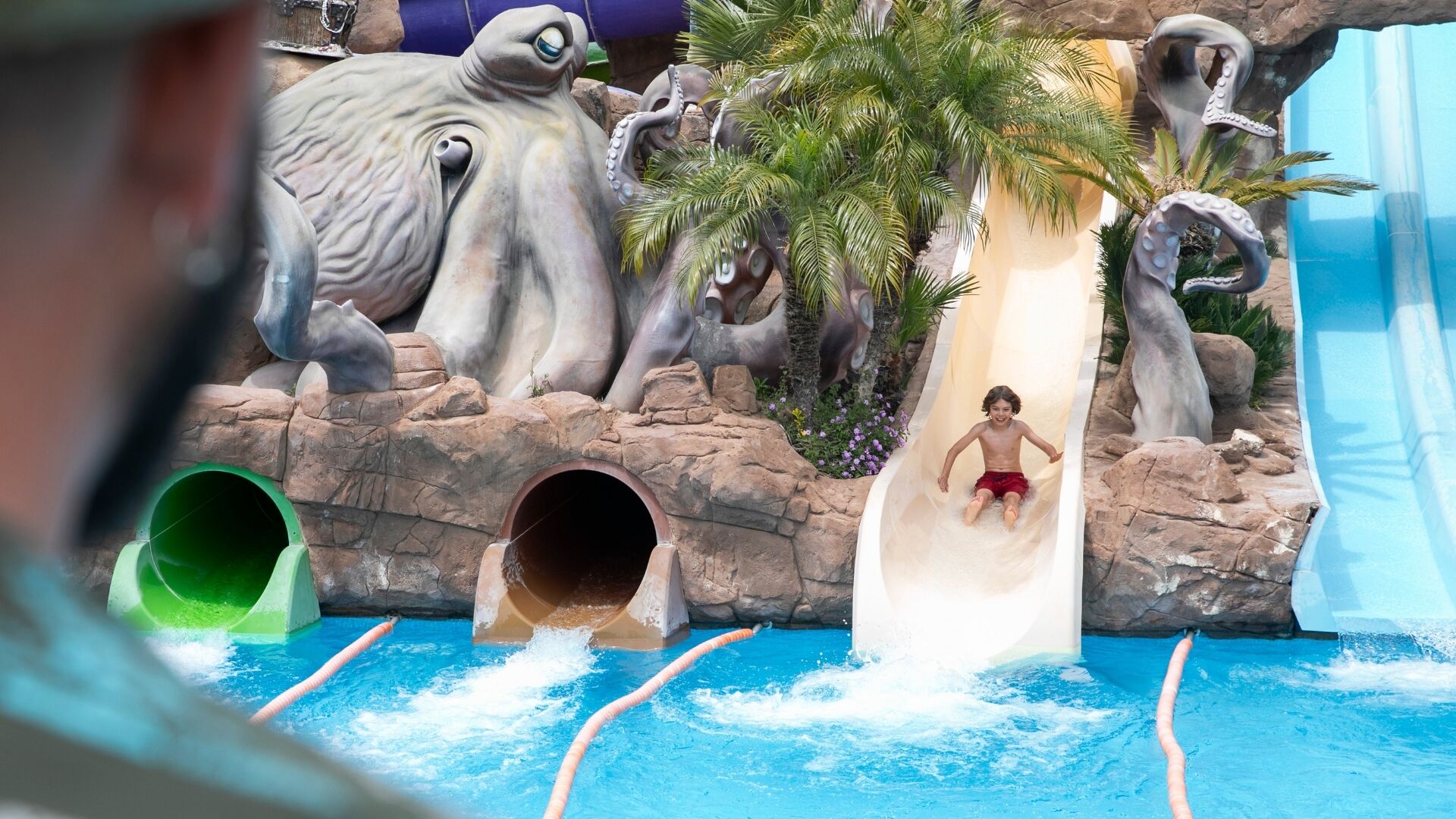A water slide with a child at the bottom, surrounded by rock formations and a pool.