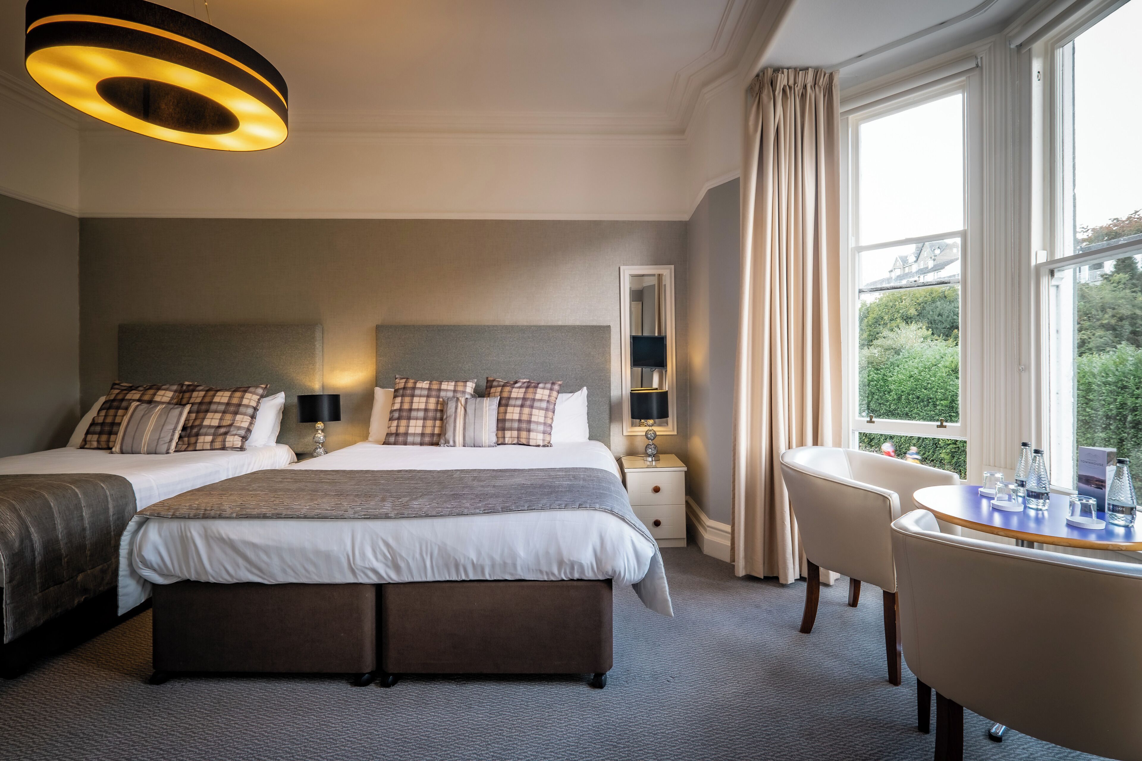 A hotel room with two beds, a desk, and a chair. There is a window with curtains, a bedside table with a lamp, and a wall-mounted mirror.