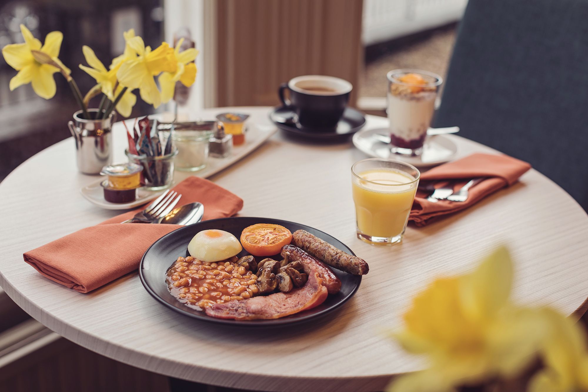 Uma mesa de café da manhã com um prato de ovos, feijão, cogumelos e linguiça, um copo de suco de laranja, uma xícara de café e um pequeno vaso com flores amarelas.