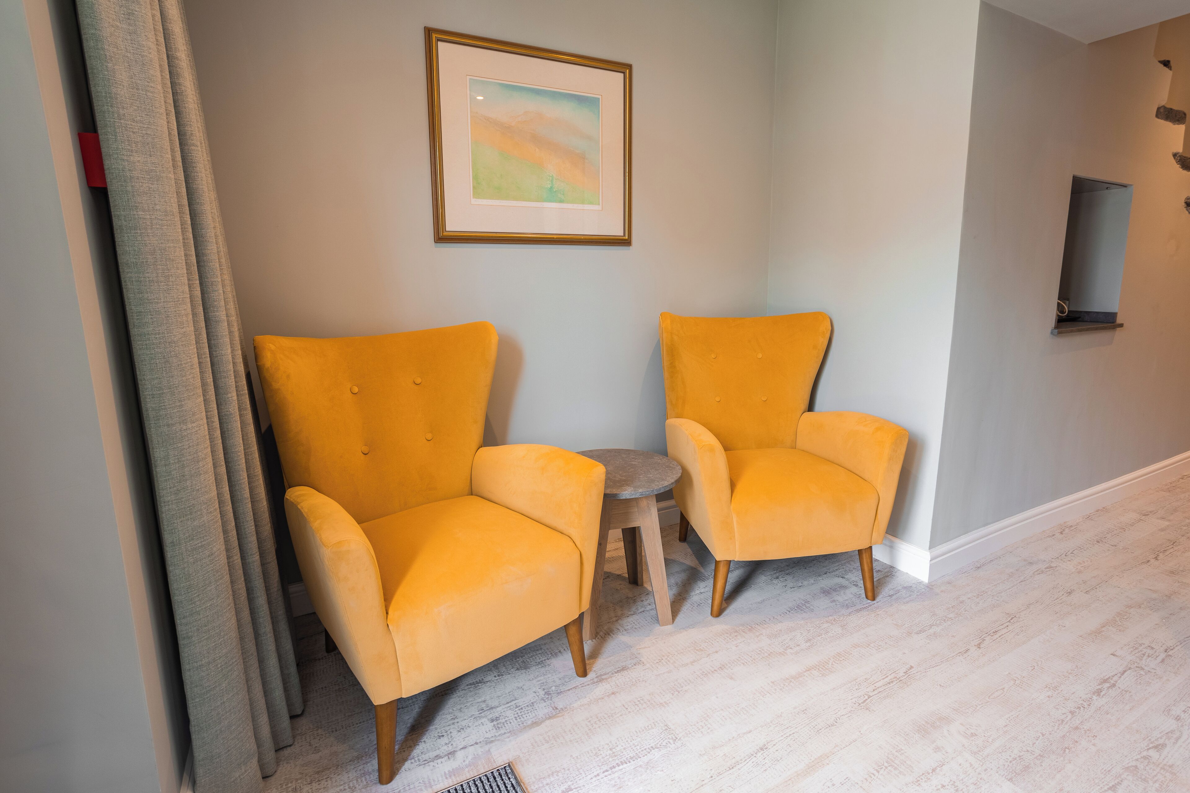 Un couloir avec deux fauteuils jaunes et une petite table ronde, un tableau encadré au mur et un plancher de couleur claire.