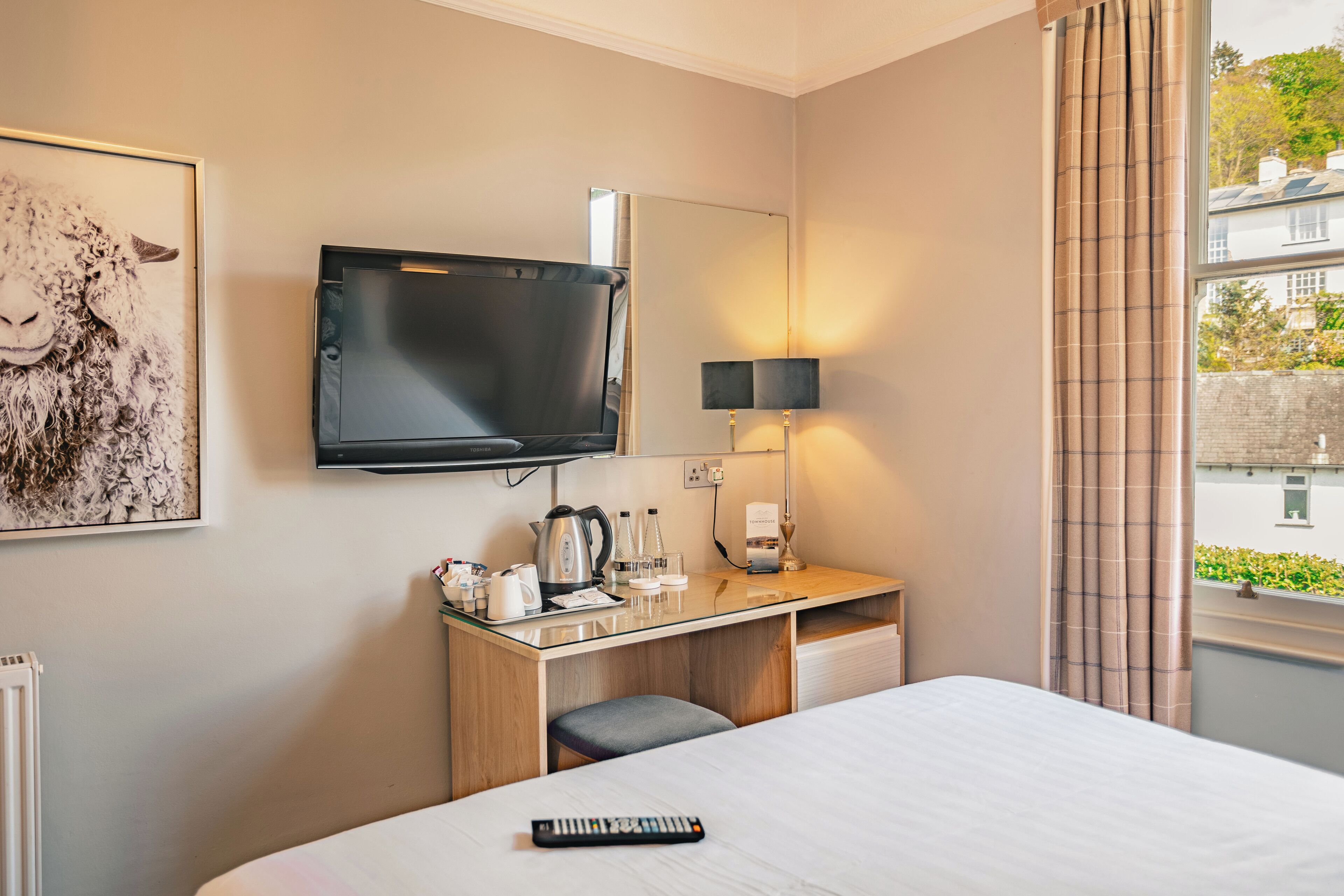 Quarto de hotel com cama, secretária com chaleira, televisão, luminária e janela com cortinas.