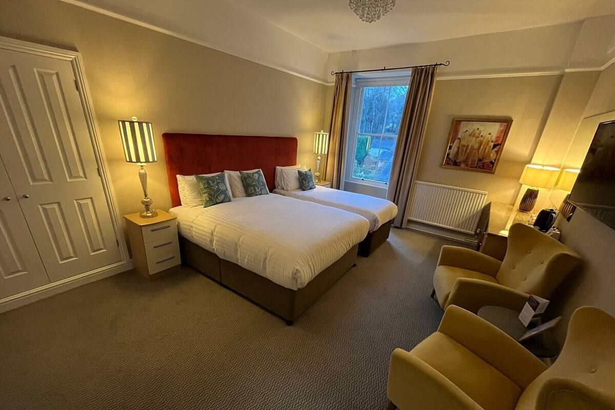 A hotel room with a large bed, two bedside tables with lamps, a chair, a window with curtains, and a radiator.