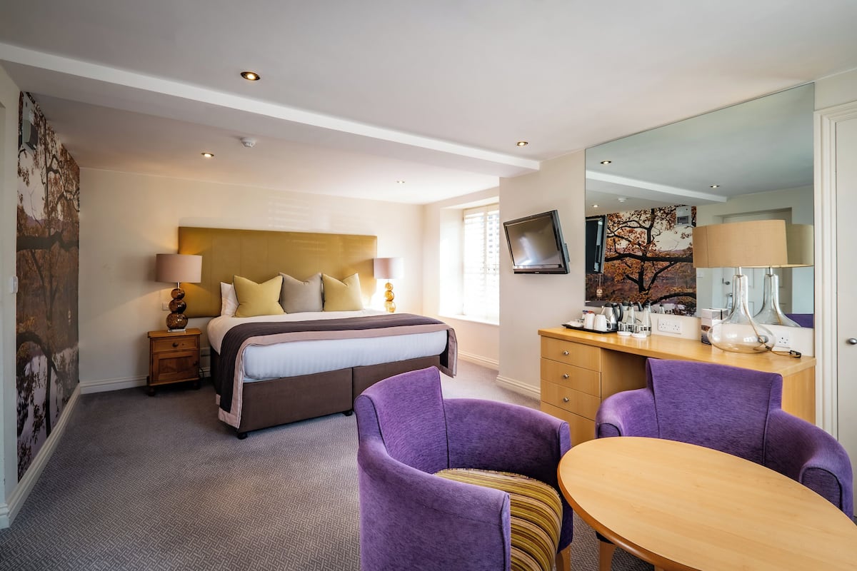A hotel room with a bed, a desk, two purple chairs, a wooden table, and a large mirror.