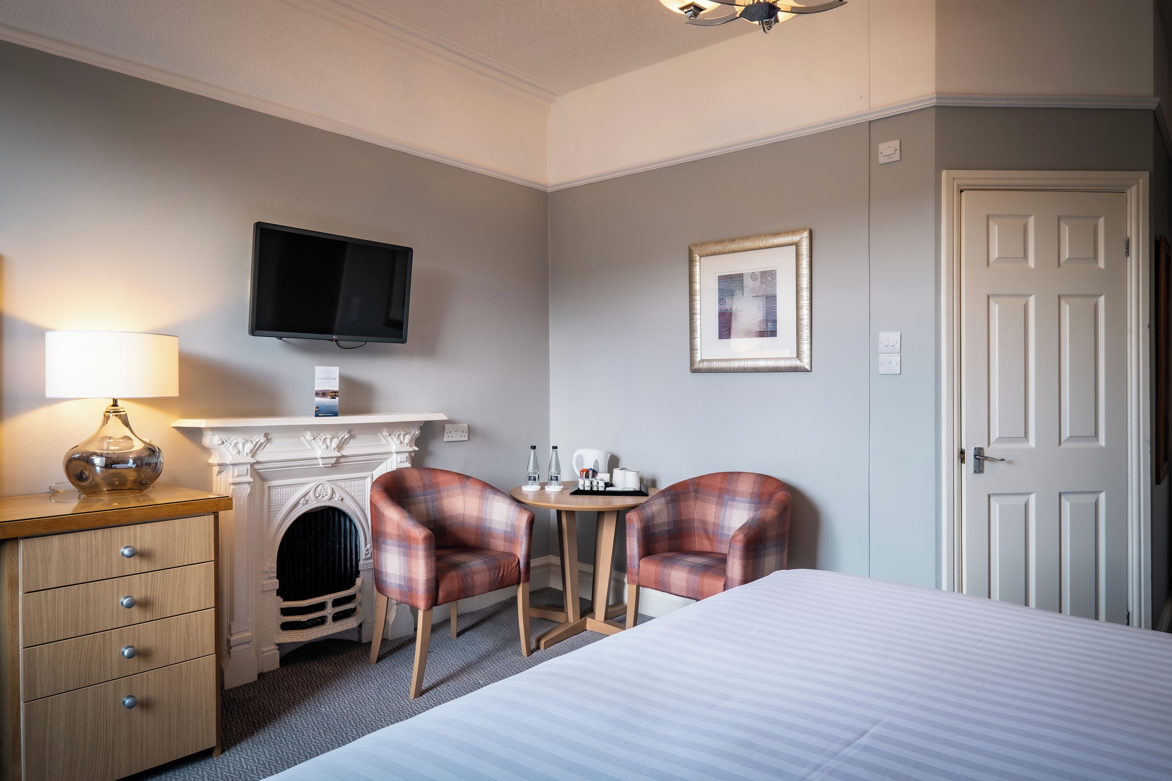 A hotel room with a bed, a desk with a lamp, two plaid chairs, a fireplace, a television, and a door.