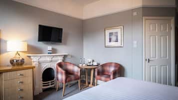 Een hotelkamer met een bed, een bureau met lamp, twee geruite fauteuils, een open haard, een televisie en een deur.