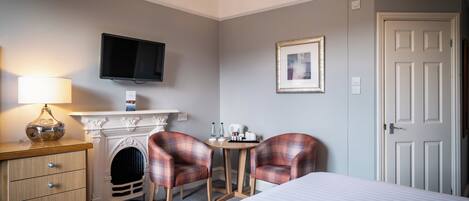 Una habitación de hotel con una cama, un escritorio con lámpara, dos sillones a cuadros, una chimenea, un televisor y una puerta.