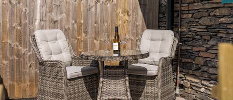 Un espacio de estar al aire libre con muebles de mimbre, dos sillas, una mesa, una botella de vino y un muro de piedra de fondo.