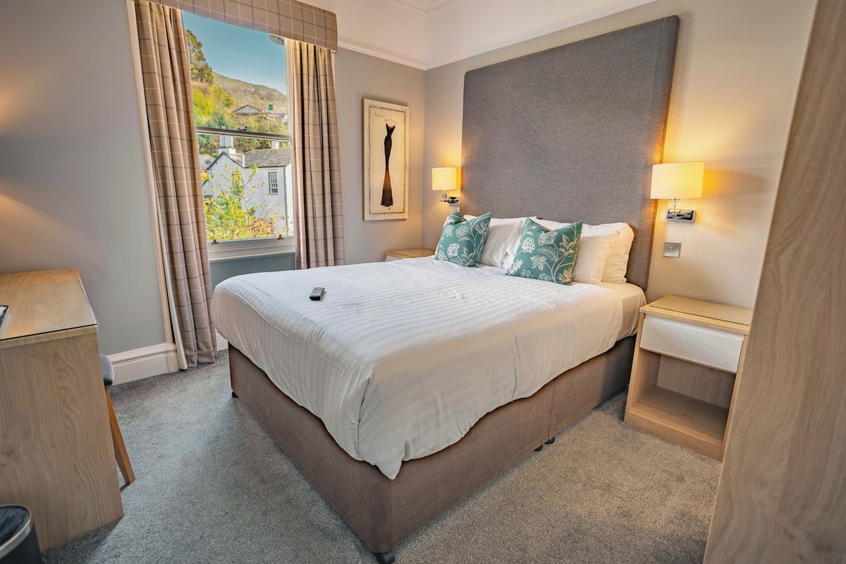 A neatly arranged hotel room with a large bed, two bedside tables, a nightstand, a window with curtains, and a framed picture on the wall.