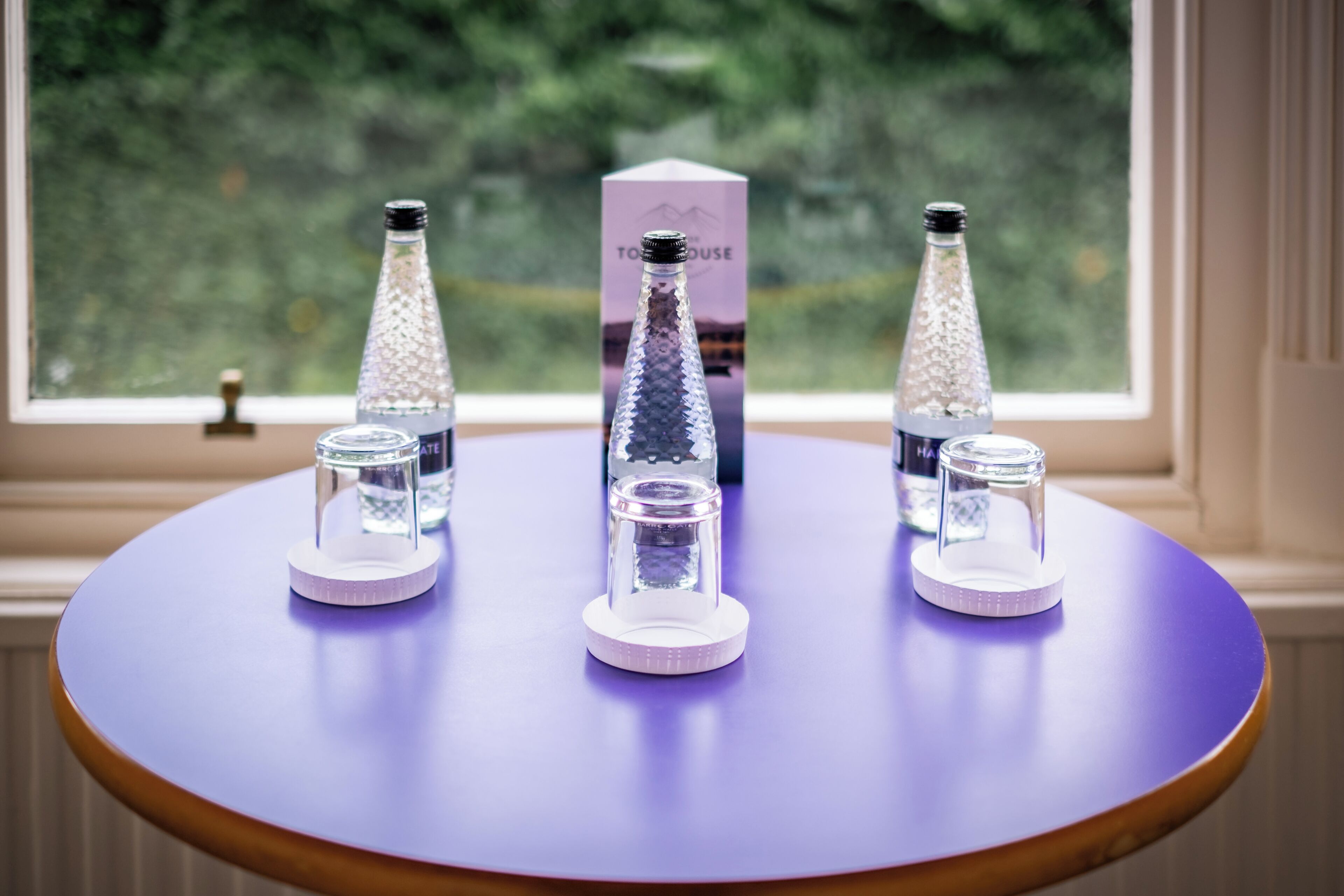 A table with three bottles of water and a box on a purple tablecloth by a window.