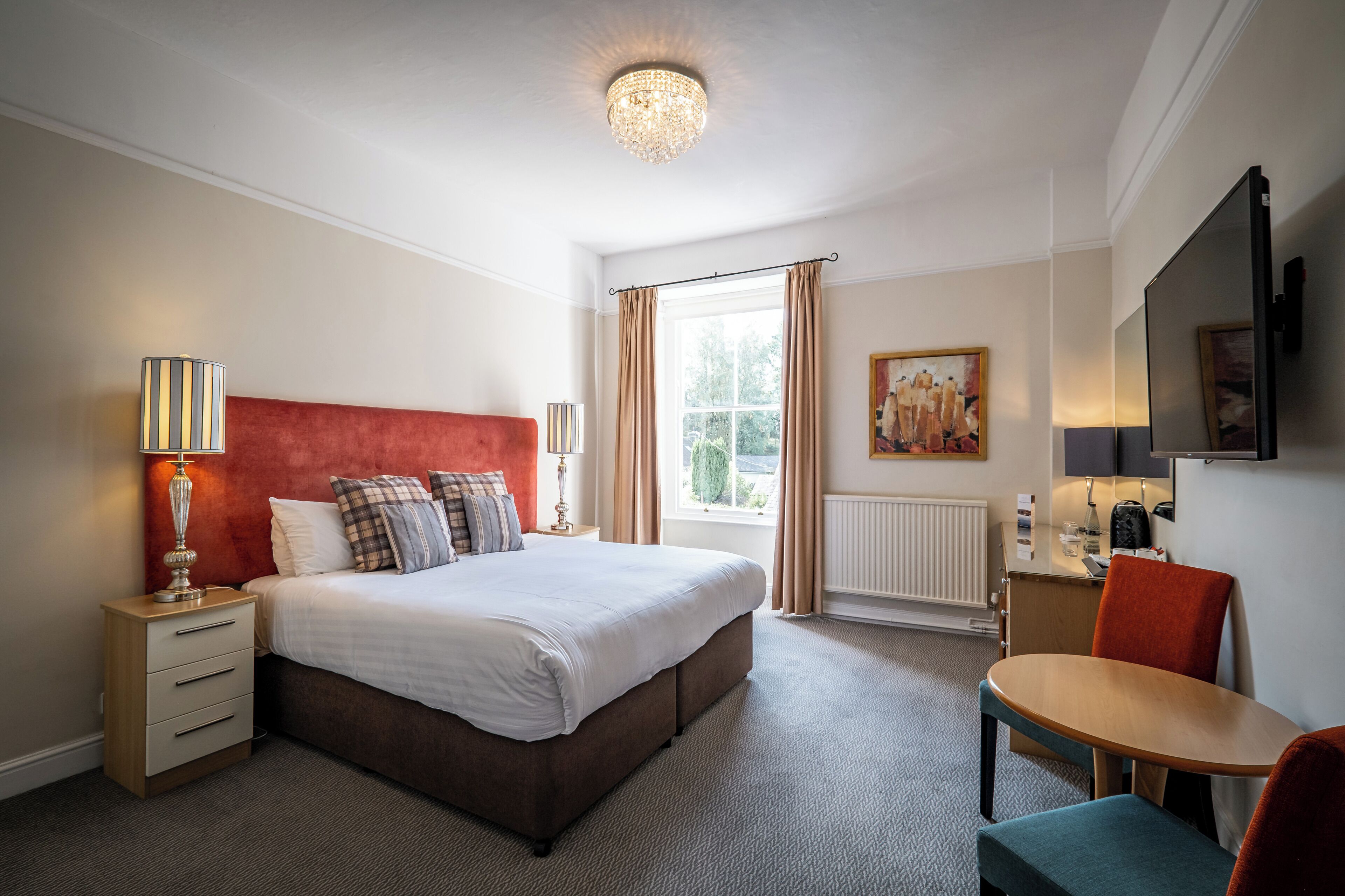 A hotel room with a large bed, a bedside table with a lamp, a small table with a chair, a window with curtains, and a painting on the wall.