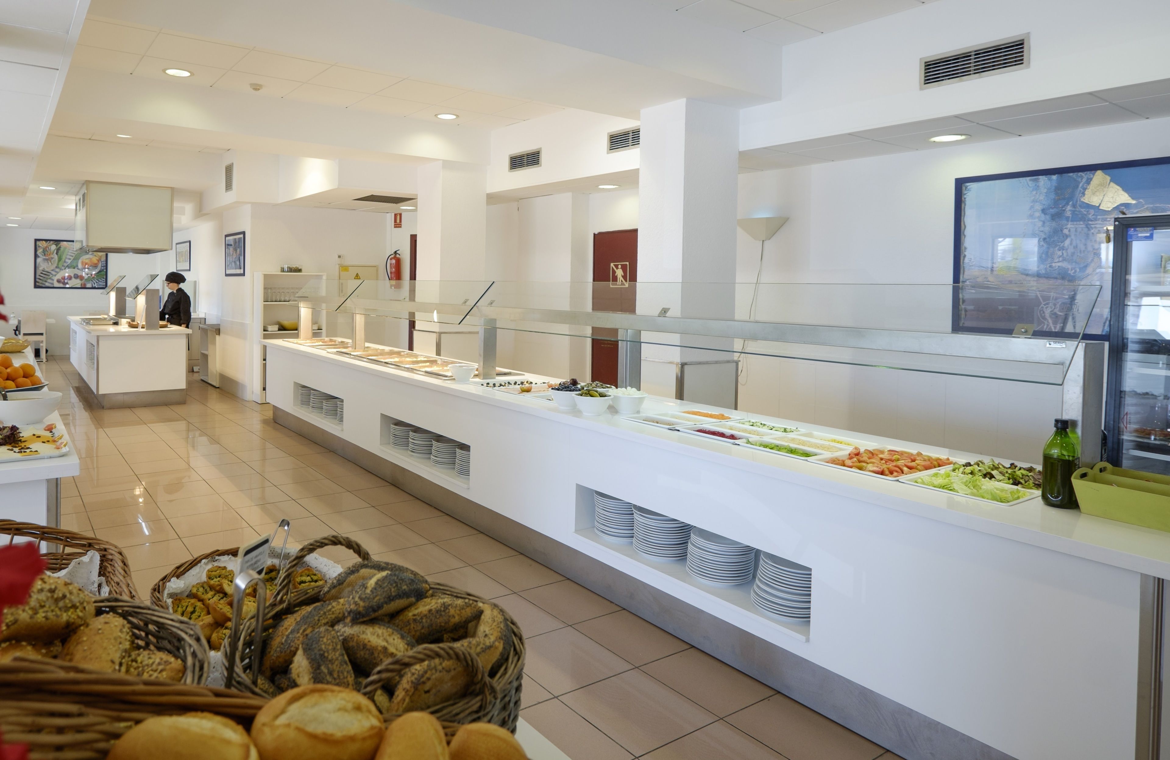 Un espace buffet avec différents stands de nourriture, une personne qui sert, et des paniers de pain.