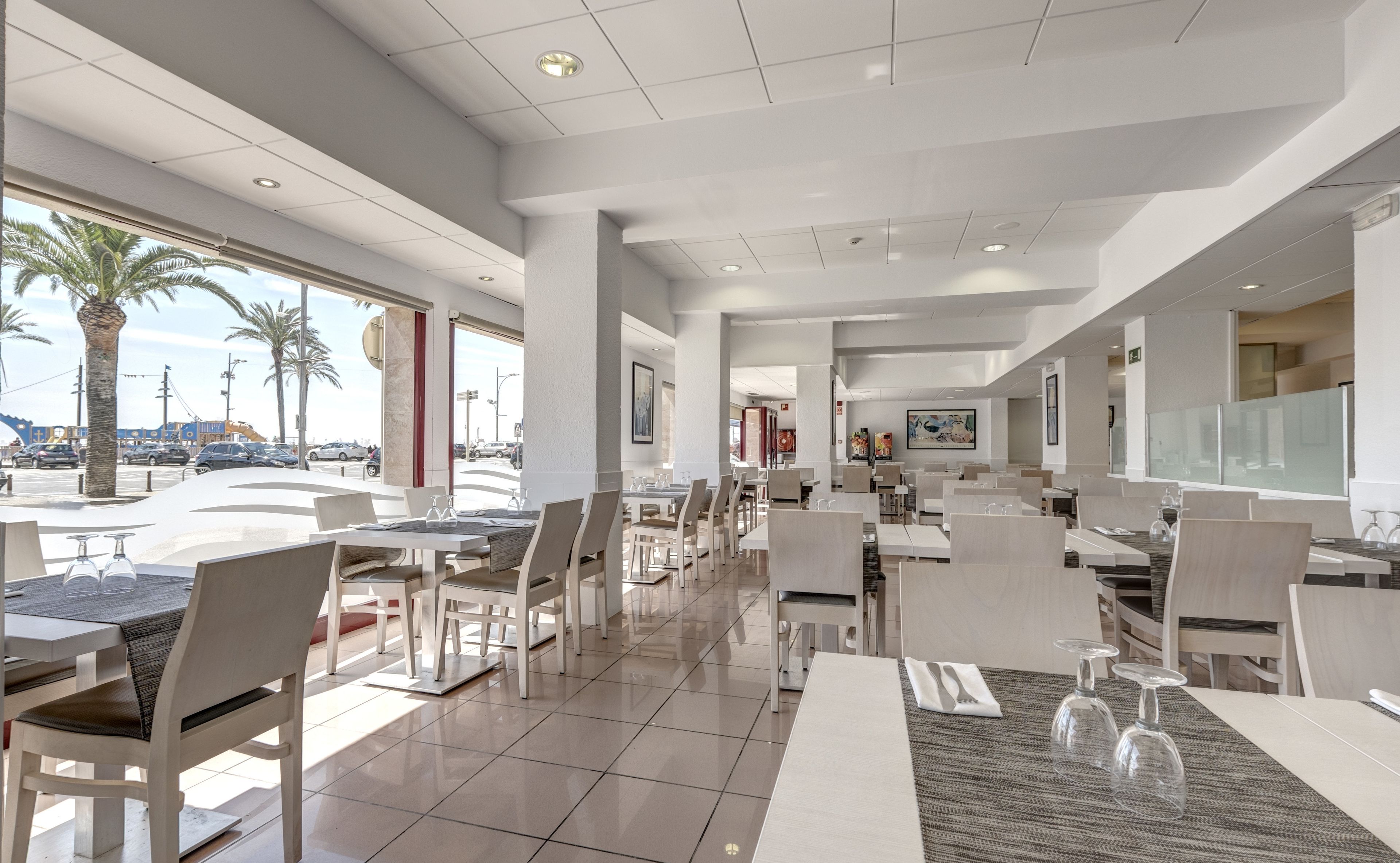 A modern restaurant with a spacious layout, large windows offering views of palm trees and a street, and a clean, contemporary design.