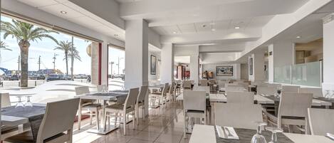 A modern restaurant with a spacious layout, large windows offering views of palm trees and a street, and a clean, contemporary design.