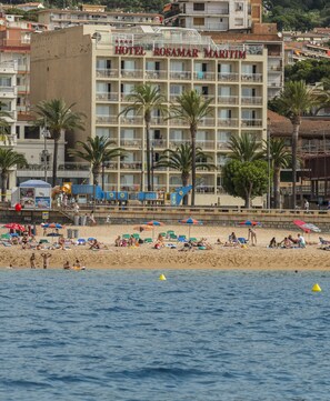 A hotel with multiple floors, balconies, and a beach area with umbrellas and people.