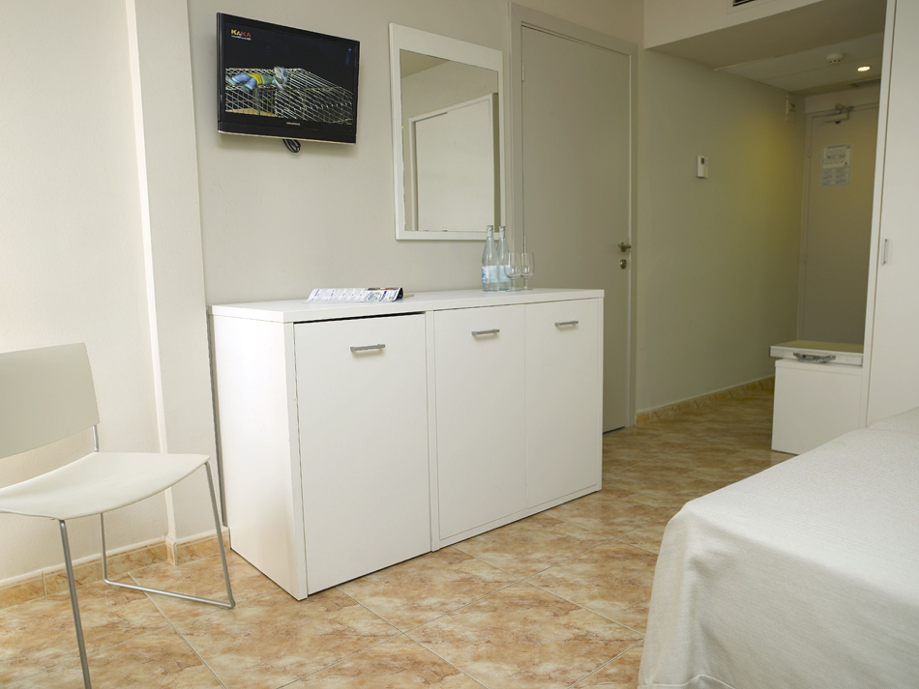 A hotel room with a white wardrobe, a television mounted on the wall, a white chair, and a bed with a white cover.