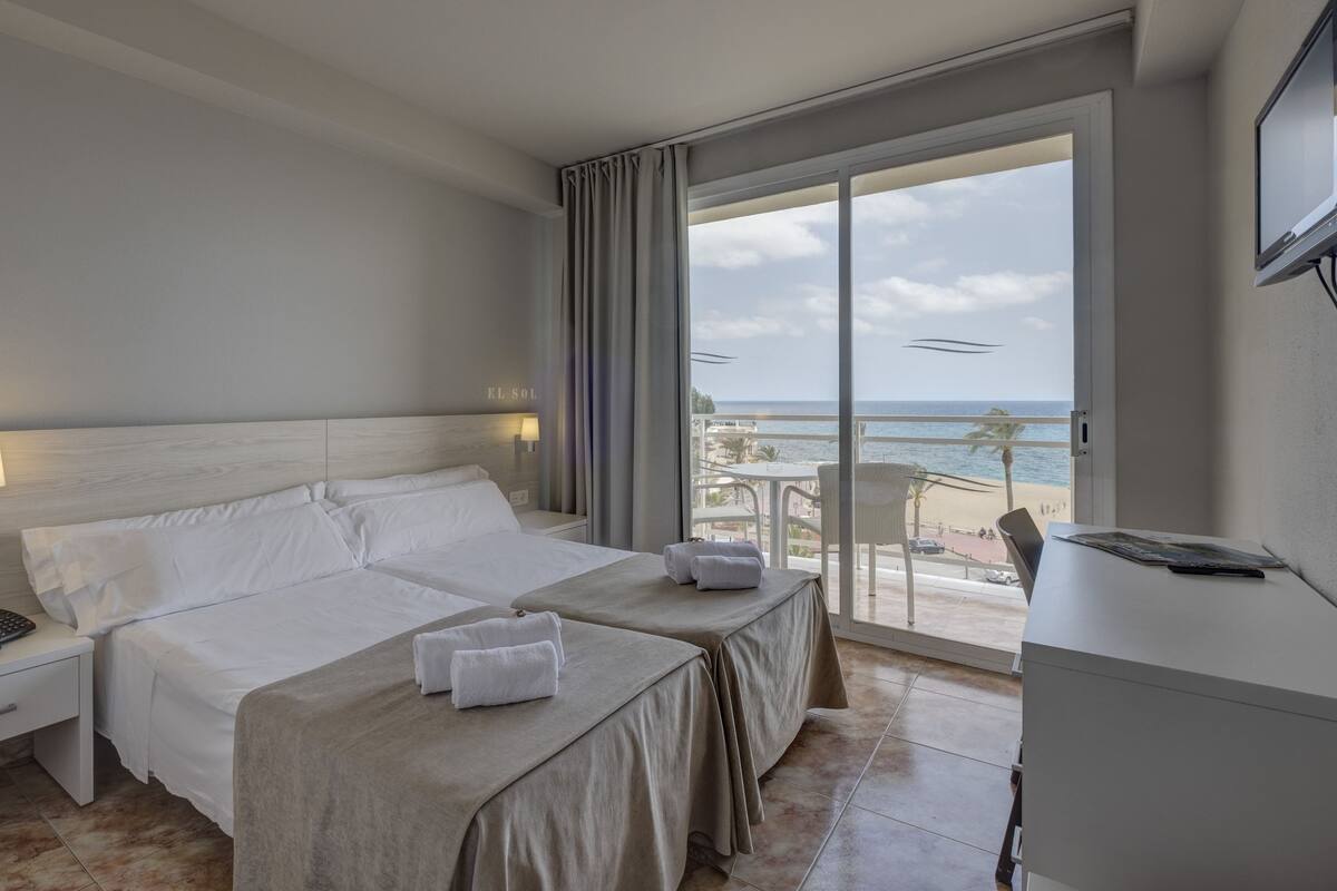 A hotel room with a large bed, two bedside tables, a desk, and a balcony with a view of the beach and ocean.