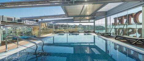 A modern indoor swimming pool with clear glass walls, metal railings, and a view of the cityscape.