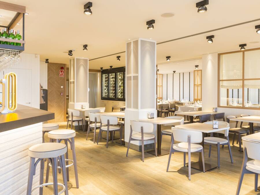 Ein modernes Restaurant mit Holzboden, weißen Tischen und Stühlen sowie einer Bar mit Sitzgelegenheiten.