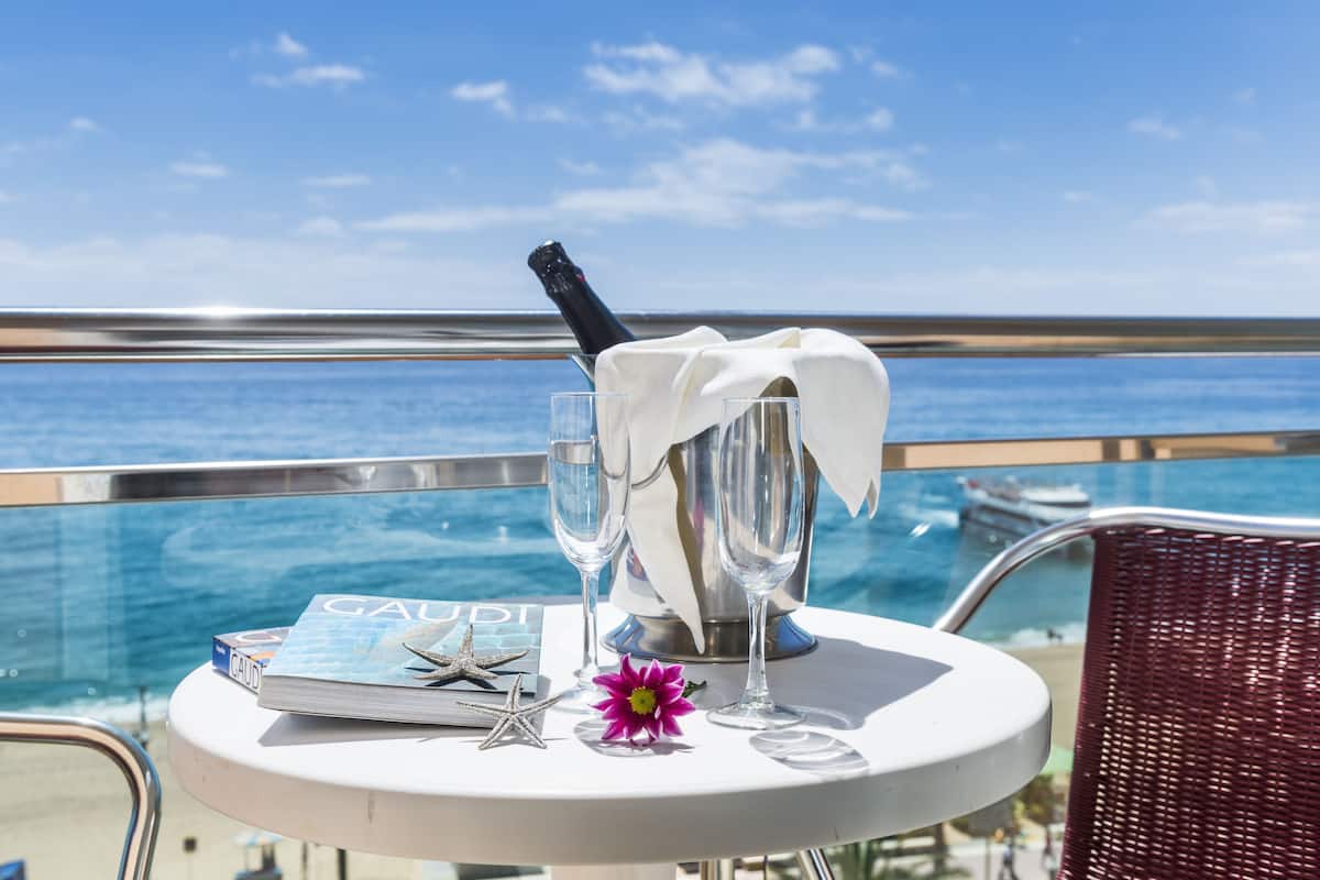 A table with a bottle of champagne, two glasses, a book titled 'GAUDI', a starfish, and a flower on a balcony overlooking the sea.