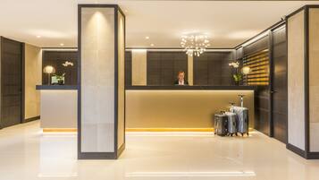 A modern reception area with a reception desk, a large pillar, and a chandelier. There are potted plants and a luggage trolley visible.