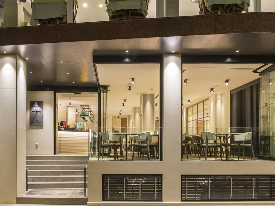 A modern building with a restaurant named 'Marsol Bar Restaurant' on the ground floor, featuring a glass facade, outdoor seating area, and a staircase leading to the entrance.