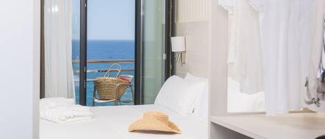 A hotel room with a bed, a suitcase, a straw hat, and a view of the sea through a large window.