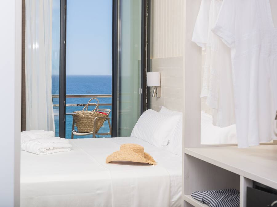 A hotel room with a bed, a suitcase, a straw hat, and a view of the sea through a large window.