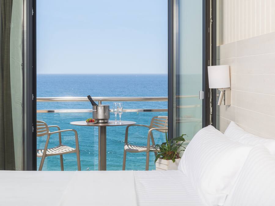 Un balcón con vista al océano, una mesa con un cubo y vasos, dos sillas y una cama con ropa blanca.